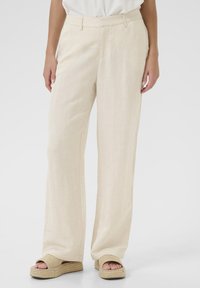 Pantalons en lin à jambe large de couleur beige clair, présentant une texture douce, un devant plat et des détails de plis subtils, associés à des sandales espadrilles beiges.