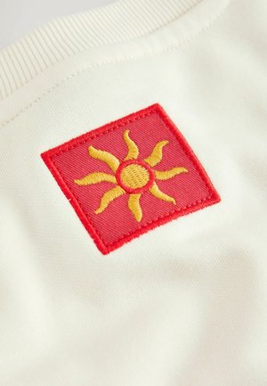 Crvena kvadratna zakrpa sa žutim izvezenim dizajnom sunca prišivena na bijelu tkaninu odjeće.