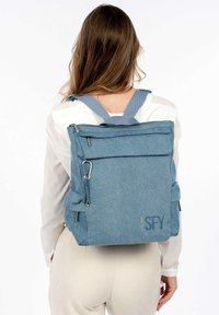 Blå tygryggsäck med en texturerad yta, rektangulär form, dragkedja upptill, justerbara remmar och sidofickor. Har logotypdetalj.