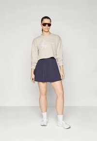 Žena u bež kratkom dukserici, tamnoplavoj suknji, bijelim čarapama, bijelim tenisicama i velikim smešnim smešnim naočalama stoji ispred jednostavne pozadine.