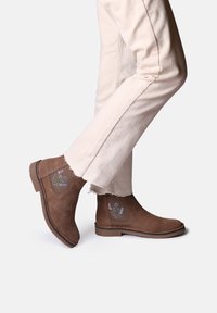 Botas de ante marrón hasta el tobillo con acentos florales bordados, tacón bajo y paneles laterales elásticos, combinadas con pantalones de color crema.