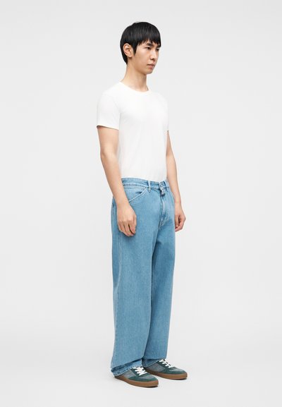 Ελαφρύ λευκό t-shirt, άνετο τζιν παντελόνι σε ανοιχτό μπλε με κανονική γραμμή, και πράσινα αθλητικά παπούτσια με λευκά κορδόνια. Κοπή με ίσια γραμμή και κλασικό σχέδιο.