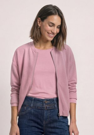 Kvinde med skulderlangt brunt hår iført en lyserød lynlåstrøje over en matchende lyserød skjorte og højtaljede blå jeans, smilende og kigger ned.