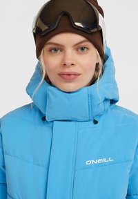 Blauwe pufferjack met een hoge kraag en een rits aan de voorkant. Bevat een logo op de borst en een gekorrelde afwerking. Draag met een bruine beanie en een bril.