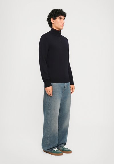 Man som bär en svart halvdraget tröja, lösa blekta blå jeans och gröna sneakers, står mot en enfärgad ljus bakgrund.