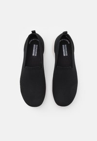 Chaussures noires à enfiler avec une tige en mesh, des panneaux latéraux élastiqués et des semelles en caoutchouc blanc. Détail du logo sur les semelles intérieures.