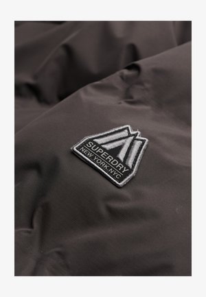 Dunkelgrauer Stoff mit einer subtilen Textur zeigt ein gesticktes Patch mit einem dreieckigen Design, auf dem "SUPERDRY" und "NEW YORK.NYC." steht.