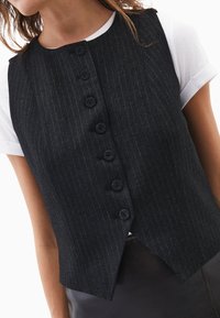 Sort vest med nålestriber, rund halsudskæring og seks store knapper. Bæres over en hvid T-shirt, og har en figursyet pasform med lige kant.