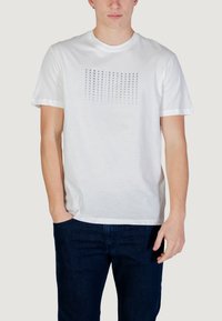 T-shirt blanc à manches courtes avec le texte "Armani Exchange" répété en dégradé de bleu, porté par une personne ayant une main dans la poche et un jean foncé.