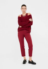 Bordeauxkleurige off-shoulder trui met lange mouwen, gecombineerd met bijpassende nauwsluitende bordeauxkleurige broek en zwarte loafers, voorzien van een decoratieve ketting.