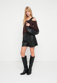 Zwarte leren shorts gecombineerd met een aansluitend bruin off-shoulder topje en zwarte kniehoge laarzen. Een zwarte schoudertas maakt de look compleet.