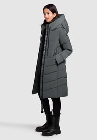 Femme portant un long manteau matelassé gris foncé avec capuche, des leggings noirs et des bottines noires, se tenant de côté avec les mains dans les poches.