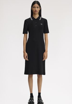 Mujer con un vestido tipo polo negro hasta la rodilla, con cuello de rayas blancas, mangas cortas, calcetines negros y mocasines negros, de pie mirando hacia adelante.