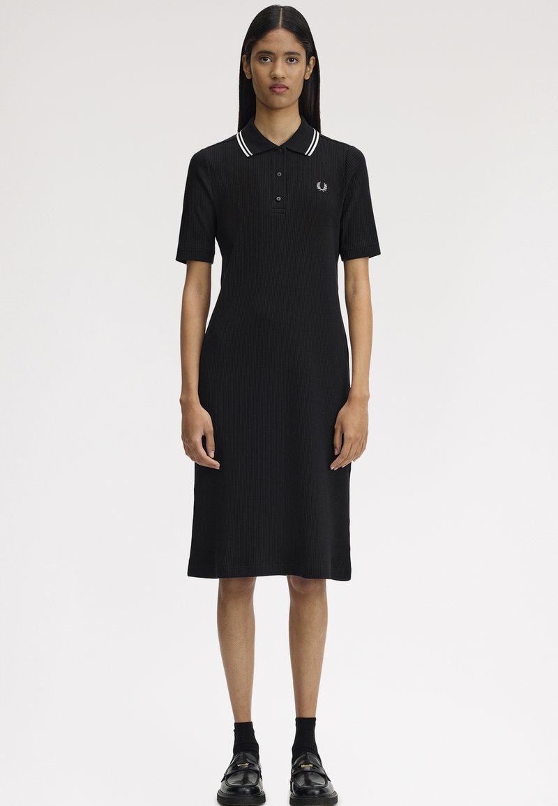 Femme portant une robe polo noire longueur genou avec un col à rayures blanches, des manches courtes, des chaussettes noires et des mocassins noirs, debout face à l'avant.