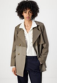 Dobbeltspent jakke i olivengrønt stoff, med bred krage, gullknapper og lagdelt design. Bæres over en hvit bluse.