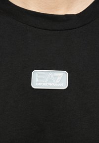 Μαύρο μπλουζάκι με στρογγυλή λαιμόκοψη και ένα μικρό ορθογώνιο ταμπελάκι με το λογότυπο EA7 Emporio Armani σε λευκό και γκρι, τοποθετημένο στο κέντρο του στήθους.