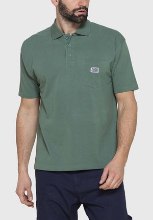 Uomo che indossa una polo verde a maniche corte con tasca sul petto e pantaloni blu navy, in piedi con le mani rilassate, guarda di lato.