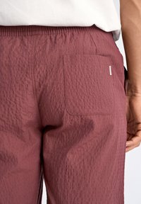 Röda byxor med textur, en bakficka och resår i midjan. Tyget har ett subtilt skrynkligt utseende.