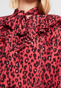 Red leopard print blouse featuring ruffled detailing along the collar, a bow tie, and a smooth, lightweight fabric texture.