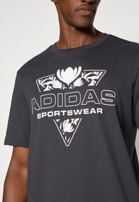 Tamsiai pilkas medvilnės marškinėliai su dideliu baltu trikampiu piešiniu, papuoštu gėlėmis, ir ryškiu užrašu "ADIDAS SPORTSWEAR".