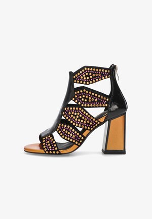 Sandalia negra de tacón alto con diseño abierto, recortes geométricos, adornos de tachuelas doradas y rosas, y un fuerte tacón cuadrado.