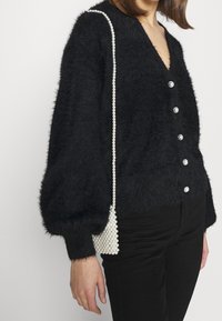 Cardigan noir et duveteux avec un col en V, six boutons argentés et des manches bouffantes volumineuses. Accompagné d'un petit sac à main orné de perles.