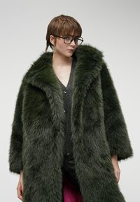 Długa, zielona sztuczna futrzana kurtka z szerokim kołnierzem, z frontem zapinanym na guziki i rękawami o długości trzech czwarte, noszona na szarym swetrze o dzianinowej fakturze.