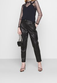 Svarta läderbyxor med lös passform, tillsammans med en genomskinlig svart långärmad topp med prickar och svarta sandaler med remmar. Liten svart handväska.