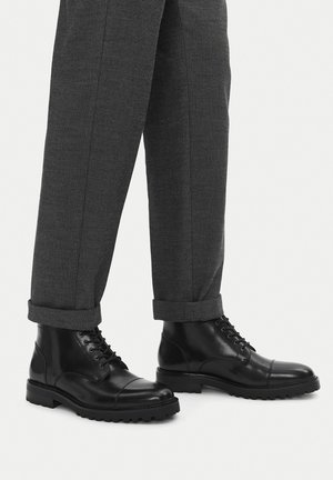 Czarne skórzane botki za kostkę o zaokrąglonym nosku, z przodu sznurowane, z wytrzymałą podeszwą; noszone z szarymi teksturowanymi spodniami podwiniętymi u dołu.