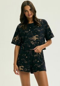 Kobieta ubrana w czarną bluzkę z krótkim rękawem i szorty, ozdobioną złotymi wzorami z Pegazem, księżycem, chmurami i gwiazdami, stoi na jednolitym tle.