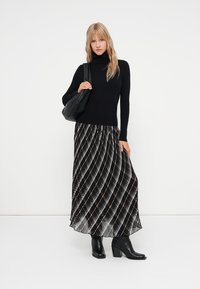 Mujer rubia vestida con un cuello de tortuga negro, falda maxi plisada en blanco y negro, botas negras hasta el tobillo, sosteniendo un bolso negro contra un fondo liso.