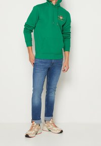 Vihreä huppari, jossa on etutasku ja kiristysnauhat, yhdistettynä sinisiin farkkuihin ja beigeihin urheilukengiin, joissa on oransseja ja vihreitä yksityiskohtia.