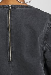 Svart denimtop med bakficka, strukturerad halsringning och en texturerad finish med synliga sömnadsdetaljer på axlarna.