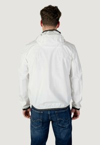 Giacca leggera bianca con cappuccio, orlo e polsini elasticizzati. Presenta una texture liscia e un design minimalista, indossata sopra jeans blu scuro.