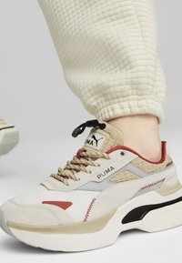 Beige und rote Puma-Sneaker mit strukturiertem Stoff, Gummisohle und gewobenen Akzenten. Mit beigen Schnürsenkeln und Logo auf der Zunge.