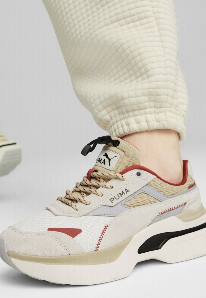 Beige und rote Puma-Sneaker mit strukturiertem Stoff, Gummisohle und gewobenen Akzenten. Mit beigen Schnürsenkeln und Logo auf der Zunge.