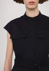 Kratka crna haljina s kratkim rukavima, mandarin ovratnikom, prednjim dugmadi, dva džepa na prsima i odgovarajućim remenom u struku.