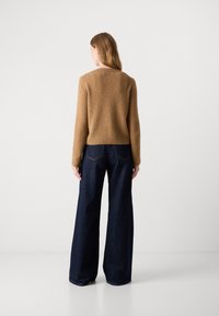 Maglione marrone con texture, a maniche lunghe, abbinato a jeans in denim scuro a gamba larga, visto da dietro su uno sfondo semplice.