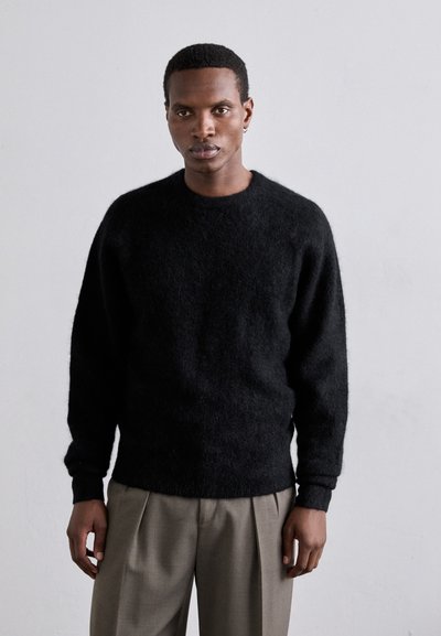 Pulover negru, pufos, din tricot, cu mâneci lungi și guler rotund, asociat cu pantaloni plisati bej deschis. Design simplu, fără modele vizibile.