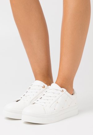 Baskets blanches basses avec côtés matelassés et petites clous dorés, portées sur des jambes nues sur un fond blanc uni.