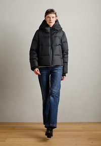 Veste doudoune noire avec capuche, design matelassé, fermeture éclair à l'avant, portée sur un jean bleu évasé et des bottines noires, le tout sur fond neutre.