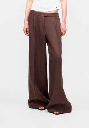 Personne portant un pantalon large marron foncé et des sandales noires à bout ouvert, debout devant un fond clair uni.