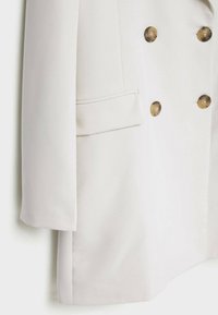 Biała marynarka podwójnie zapinana na guziki w kolorze złotym, gładki materiał, klapy w szpic oraz pojedyncza kieszeń na piersi. Cechuje się strukturalnym dopasowaniem i długimi rękawami.