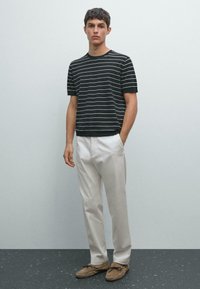 Giovane in piedi, indossa una camicia a maniche corte a righe bianche e nere, pantaloni beige chiaro e mocassini marroni.
