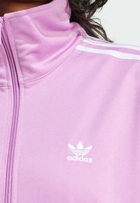 Närbild av en person som bär en ljuslila Adidas zip-up jacka med vita ränder på axeln och Adidas-logotypen på bröstet.