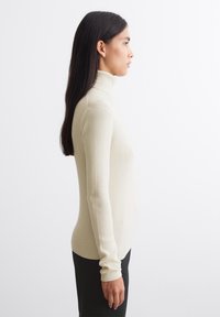 Cremfarbener, gerippter Rollkragenpullover mit langen Ärmeln, figurbetontem Design und glatter Textur. Stilisiert mit schwarzen Hosen, von der Seite gezeigt.