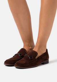 Mocassins en daim marron avec un talon plat, un bout arrondi et des détails en métal noir sur le cou-de-pied. Texture lisse avec des coutures minimales.
