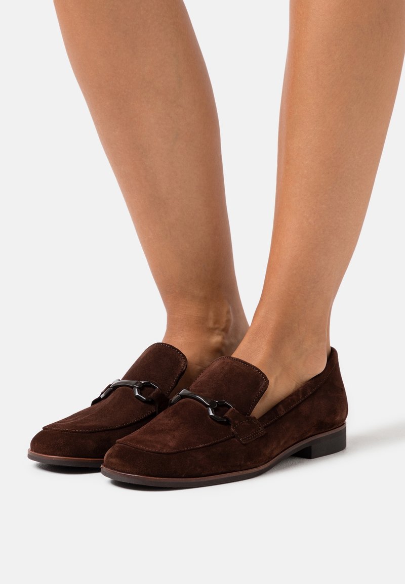 Mocassins en daim marron avec un talon plat, un bout arrondi et des détails en métal noir sur le cou-de-pied. Texture lisse avec des coutures minimales.