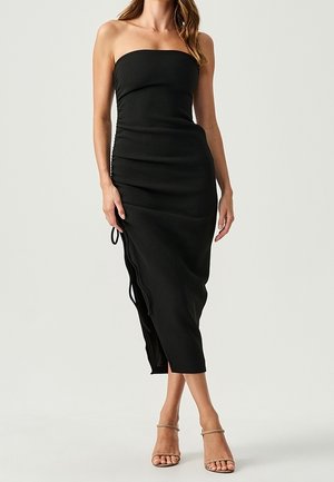 Femme portant une robe midi noire bustier avec fronces sur le côté et des sandales à talons beiges, debout devant un fond uni clair.