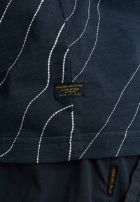Navy blue fabric featuring white wavy lines and a black tag with yellow text. The material is textured with a lightweight appearance.
